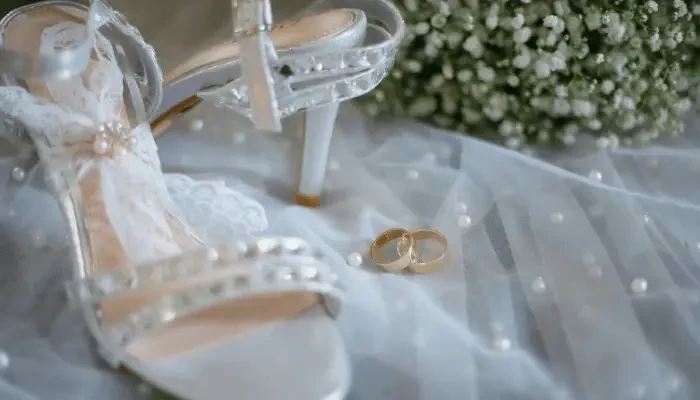 White lace bridal shoes with pearl details beside gold wedding rings on a sheer pearl-studded wedding veil.