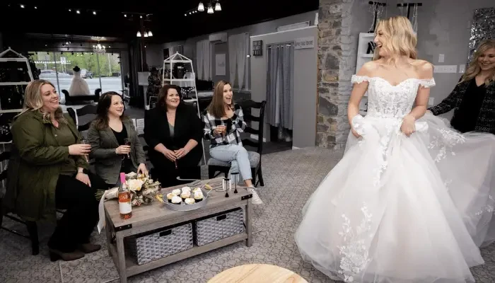 Bride wearing an off-the-shoulder lace ball gown while showing her wedding dress to friends during a bridal appointment.