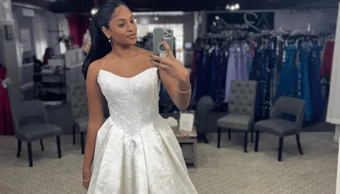 Bride taking a mirror selfie while wearing a white strapless wedding dress with a full embroidered skirt in a bridal boutique.