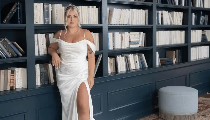 Bride in a white satin wedding dress standing in front of a bookshelf, posing for a photo.