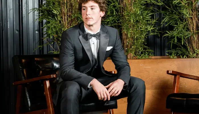 Groom in a dark tuxedo sitting in a leather chair in a stylish setting.