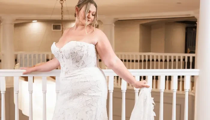 Bride in a strapless lace wedding dress standing on a balcony and holding her train.