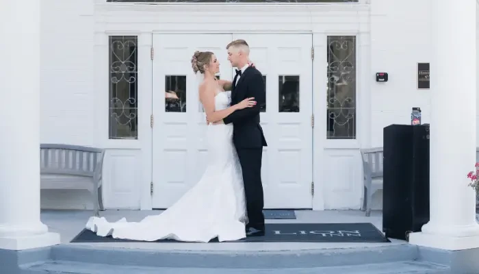 Bride and groom standing together outside a white building, with the bride wearing a fitted wedding gown with a long train and the groom wearing a black tuxedo.