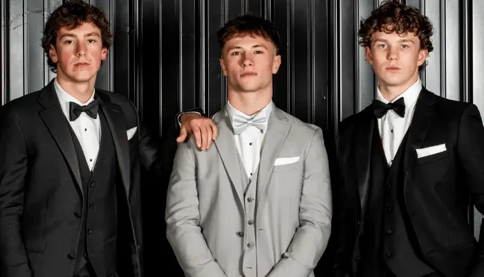 Three men posing together in formal tuxedos, with the man in the center wearing a light gray tuxedo and the men on either side wearing black tuxedos.
