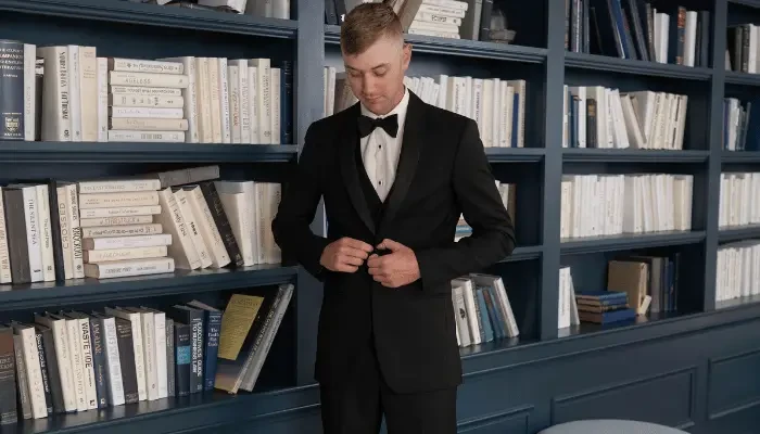 Man wearing a black tuxedo and bow tie while standing in front of dark blue bookshelves.