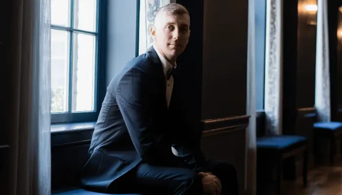 Man wearing a dark suit and white shirt sitting beside a window in an elegant interior.