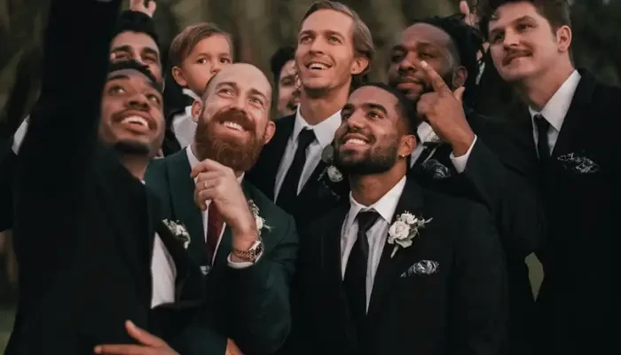 Group of men in formal suits and tuxedos gathered outdoors and looking toward a phone while taking a group selfie.