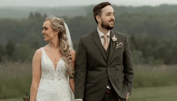 Bride and groom standing together in an outdoor field, with the bride wearing a lace wedding dress and the groom wearing a dark green suit with a boutonniere.