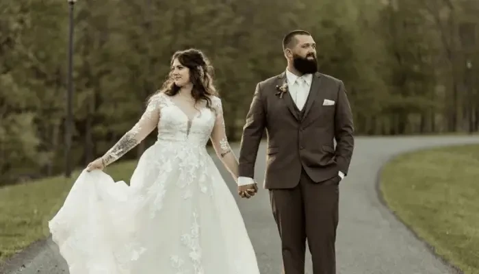 Bride and groom walking hand in hand outdoors, with the bride wearing a long-sleeved lace wedding dress and the groom wearing a brown suit.