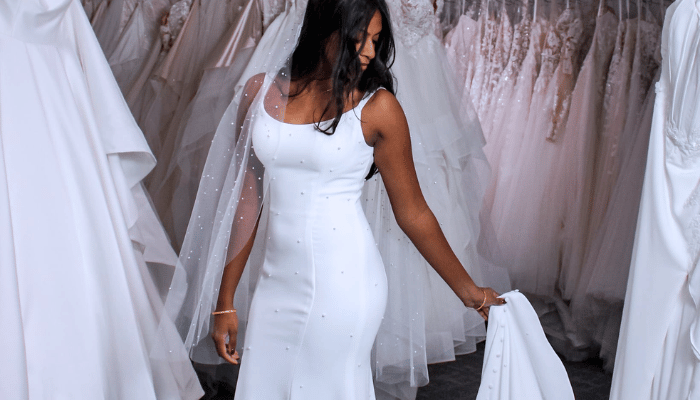 Bride in a fitted white wedding dress with a sheer veil, standing in a bridal boutique surrounded by gowns