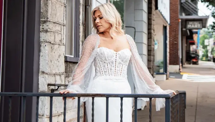 Bride in a white wedding dress with sheer sleeves standing on a balcony outside a building.
