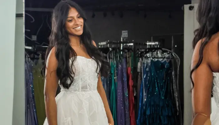 Smiling bride in a white floral wedding dress admiring her reflection in a bridal boutique mirror.