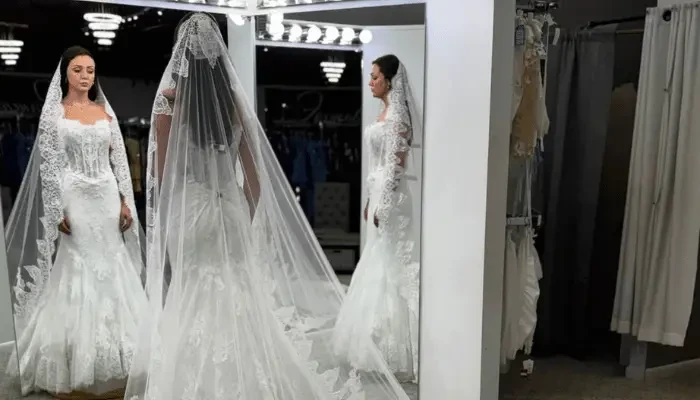 Bride in a lace wedding gown and long veil standing in front of mirrors inside a bridal boutique.