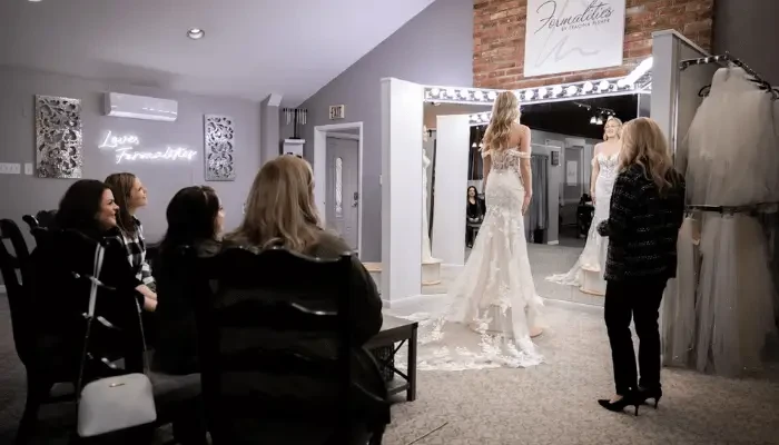Bride in a wedding gown standing before a mirror while family and a bridal consultant watch during a dress appointment at a bridal boutique.