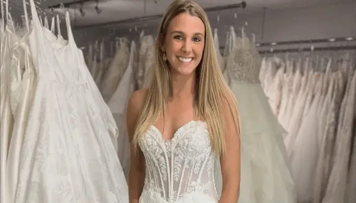 Smiling bride in a lace wedding dress standing among rows of wedding gowns in a bridal boutique.